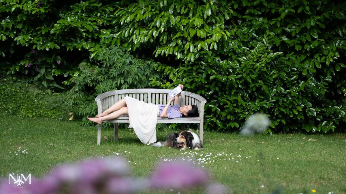 Beatrice lying on the bench reading a book in the garden while pet sitting Charlie the dog who is relaxing under the bench with his tongue sticking out