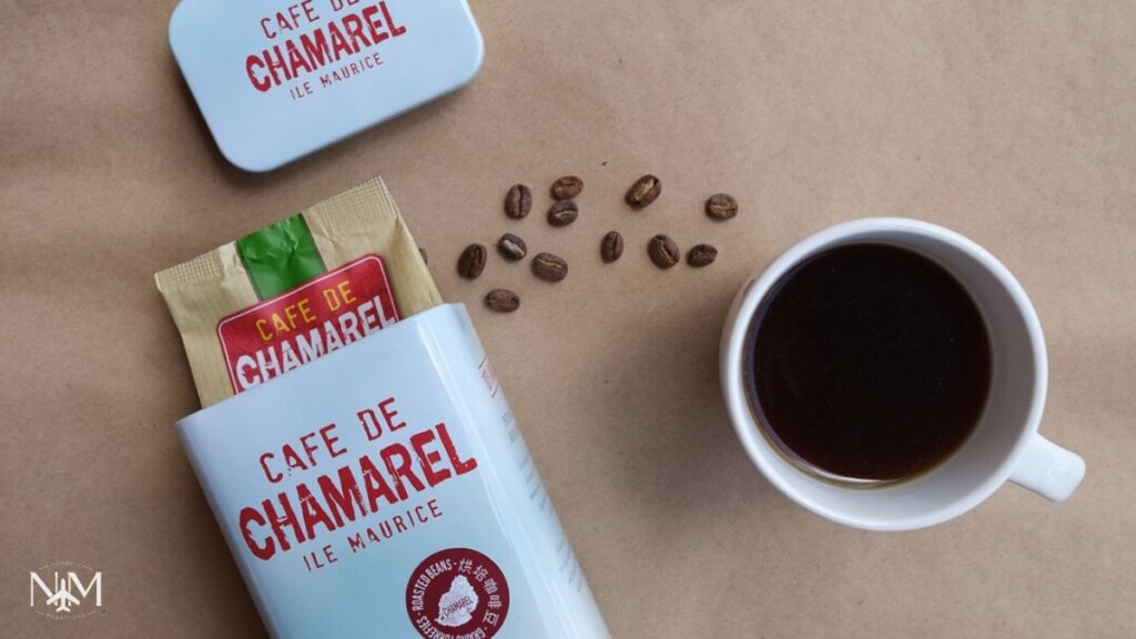 a cup of coffee and a box of freshly arabica coffee beans named Cafe de Chamarel, Ile Maurice. Some coffee beans are scattered.